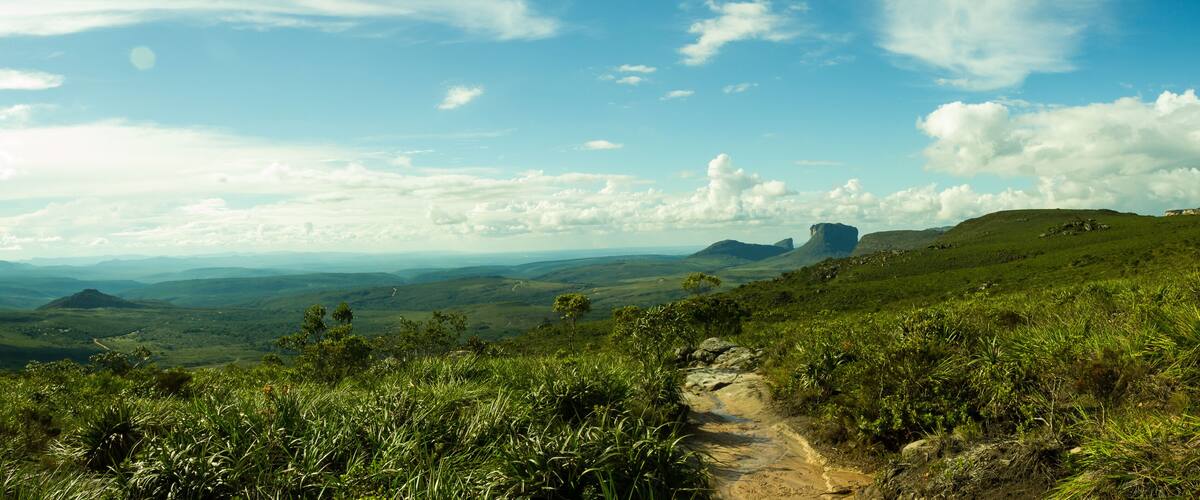 A hiking trail through a lush green landscape with rolling hills and distant mountains under a partly cloudy sky.