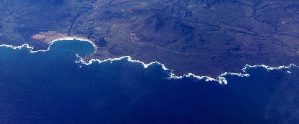 An airplane wing with a view of snow-capped mountains and a coastline below.