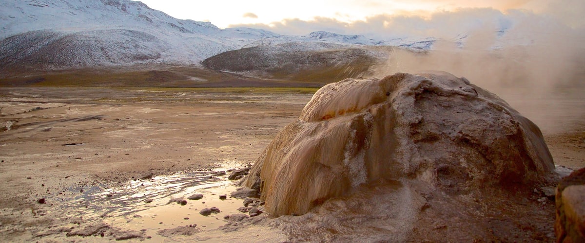 A geyser in a dry, mountainous landscape with snow-capped peaks in the background.