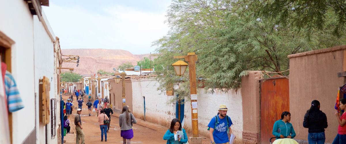 A narrow dirt road lined with buildings and trees, with people walking and a stroller visible.