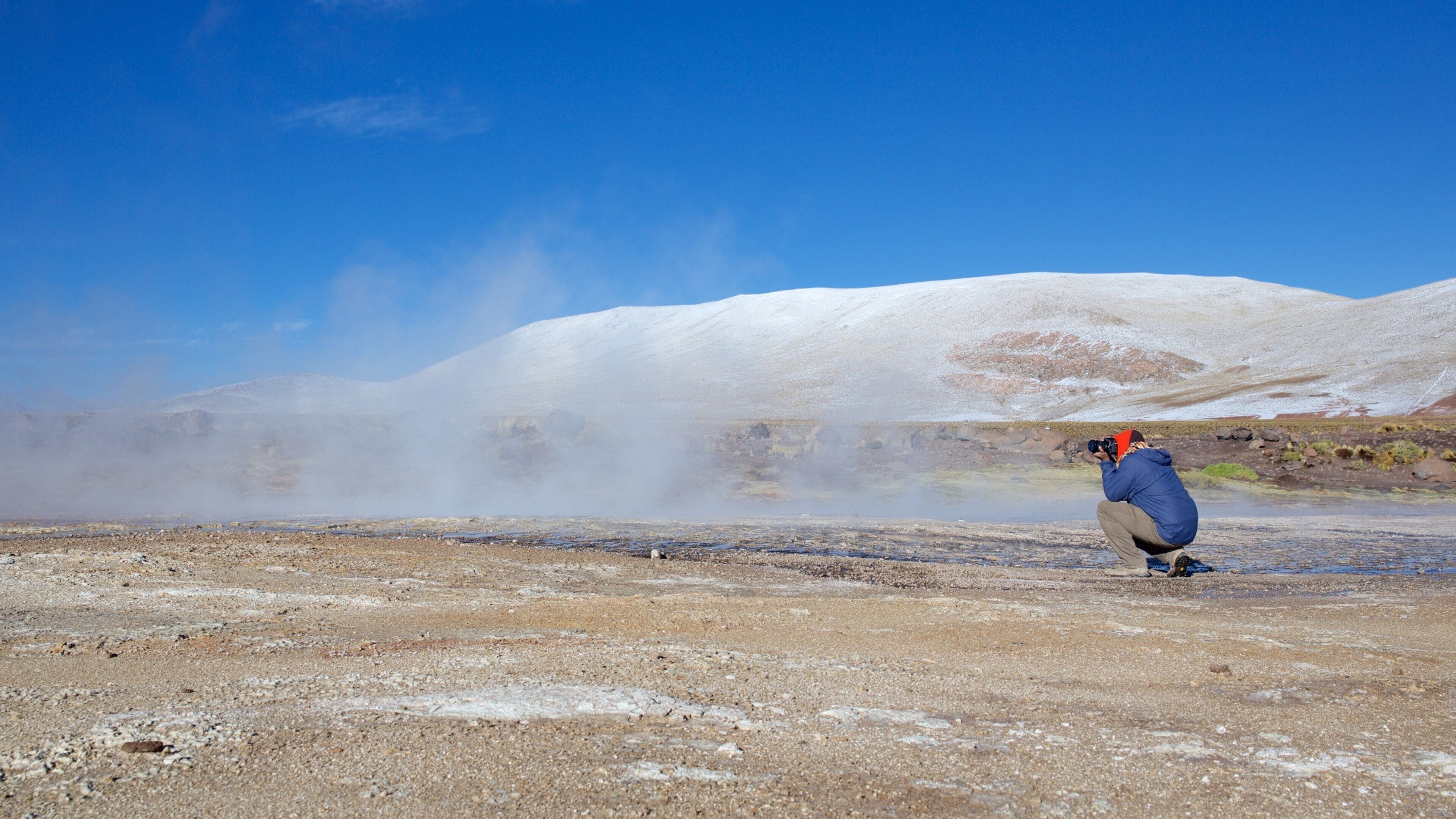 Una persona fotografiando un área geotérmica con vapor ascendiendo del suelo y colinas nevadas al fondo.