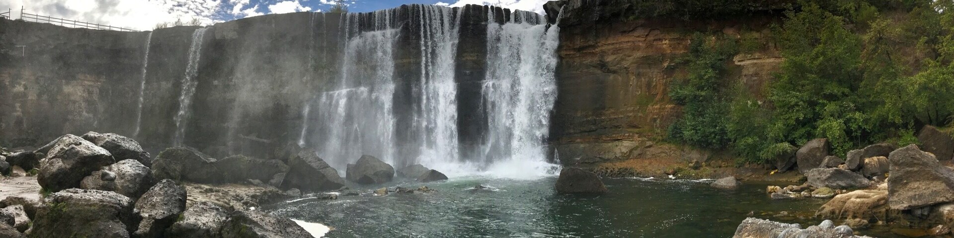 A waterfall cascading over a cliff into a river surrounded by rocks and greenery.