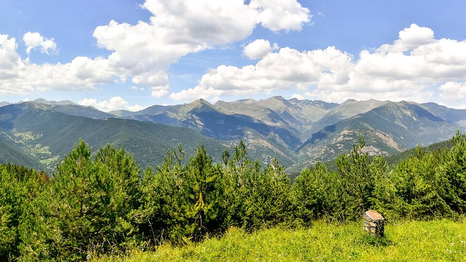 A mountainous landscape with dense green forests and a clear blue sky with scattered clouds.