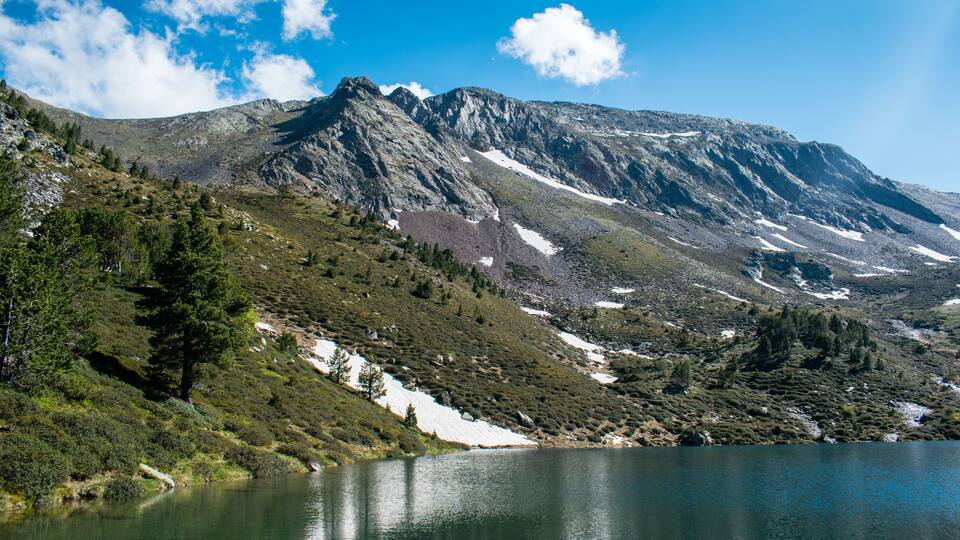 A mountainous landscape with a lake, snow patches, and dense greenery.
