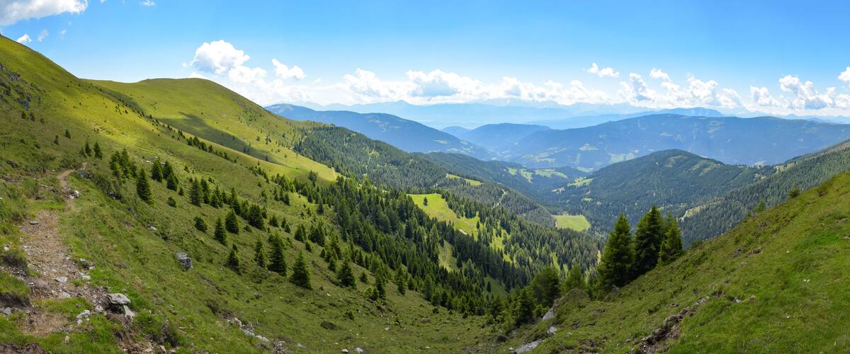 A mountainous landscape with a valley, forested areas, and distant mountain ranges under a clear sky.