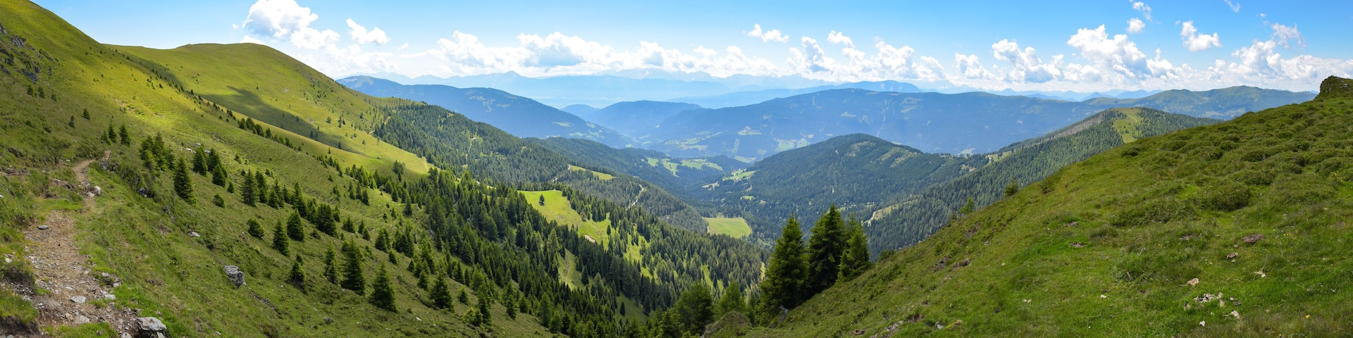 A mountainous landscape with a valley, forested areas, and distant mountain ranges under a clear sky.