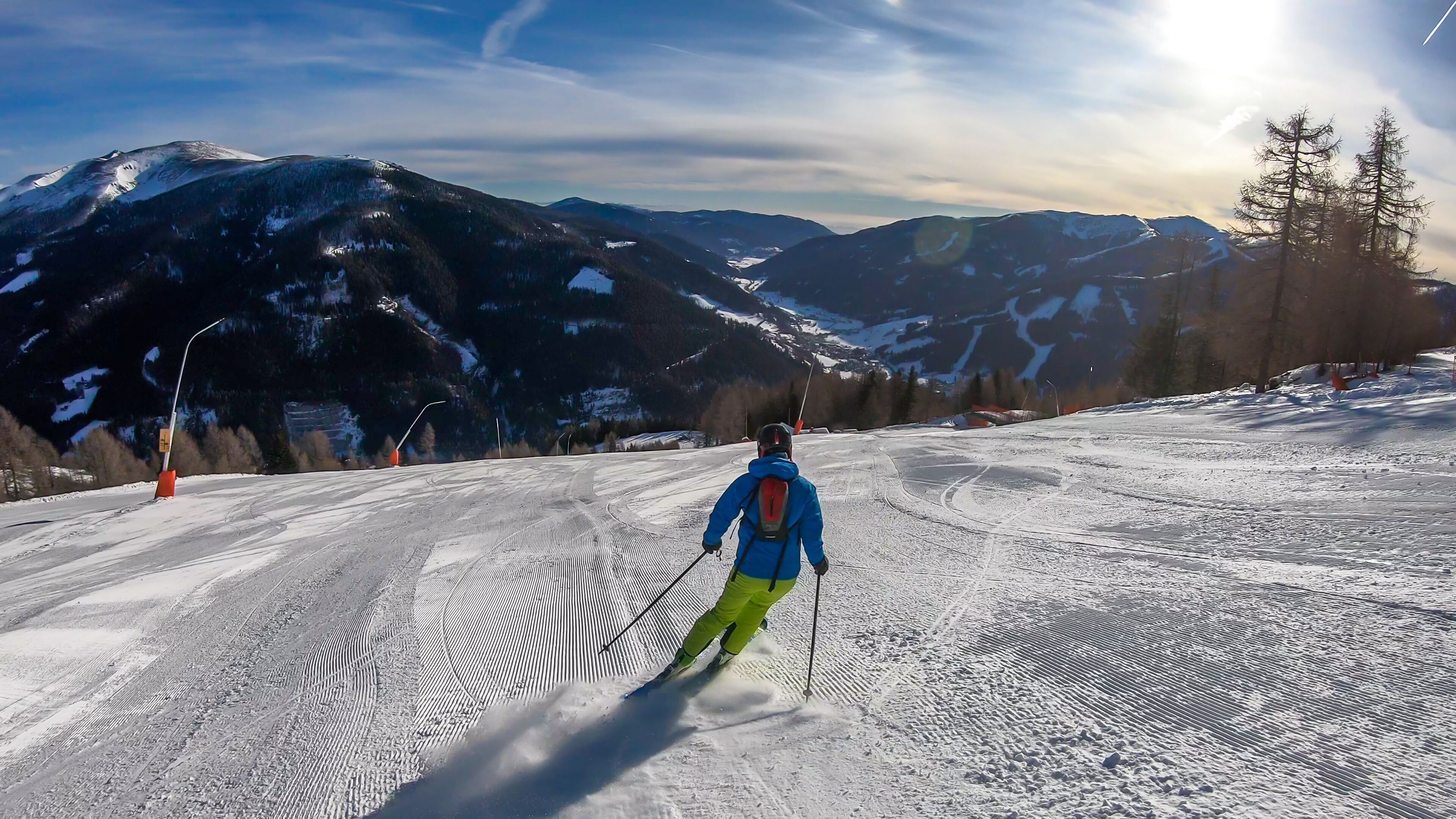 A man with a small backpack skiing on a perfectly groomed slopes in Bad Kleinkirchheim, Austria. There are lots of snow caped mountains around him. Few trees on the side. Winter sports.