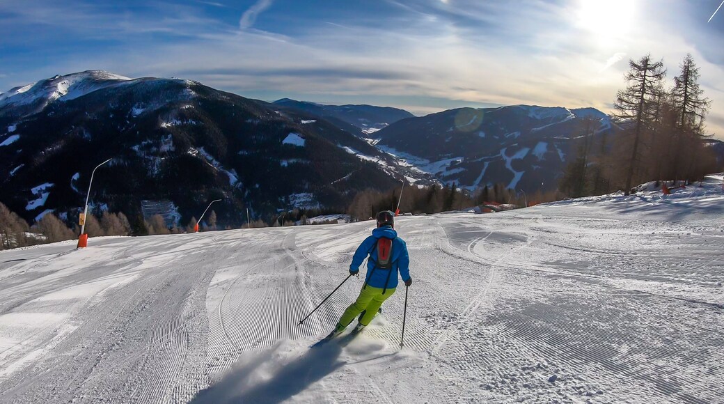 A man with a small backpack skiing on a perfectly groomed slopes in Bad Kleinkirchheim, Austria. There are lots of snow caped mountains around him. Few trees on the side. Winter sports.