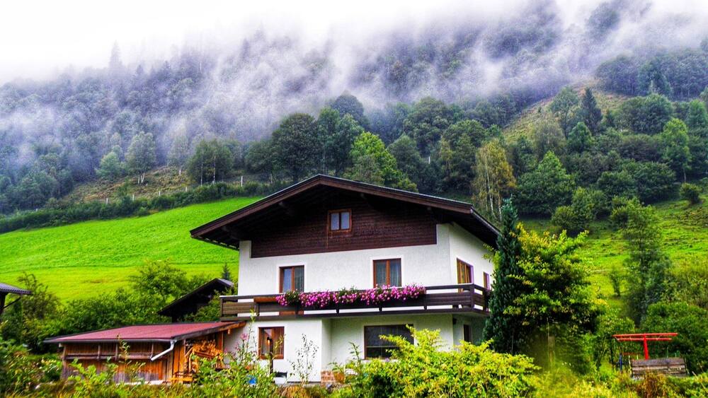 Ein zweistöckiges Haus mit weißer Fassade und braunem Dach, umgeben von üppiger Vegetation und einem bewaldeten Hang.