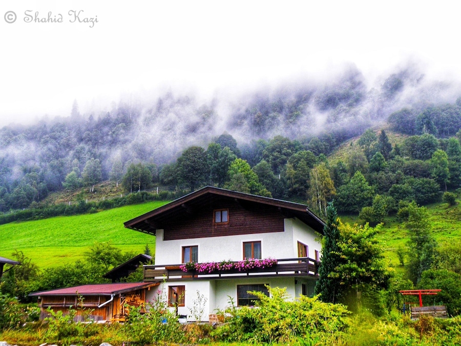 Ein zweistöckiges Haus mit weißer Fassade und braunem Dach, umgeben von üppiger Vegetation und einem bewaldeten Hang.