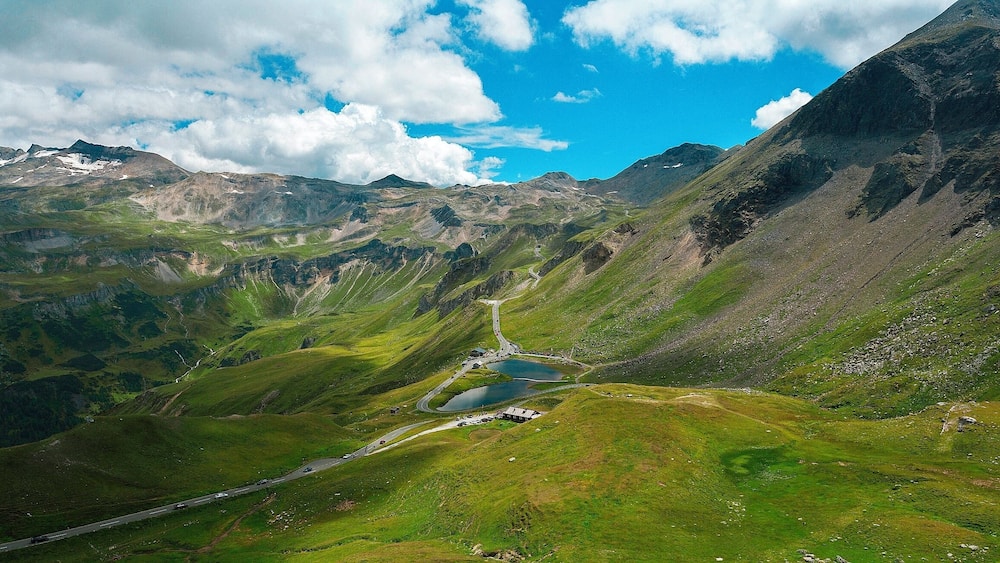 A mountainous landscape with a winding road, a small lake, and a building near the road.