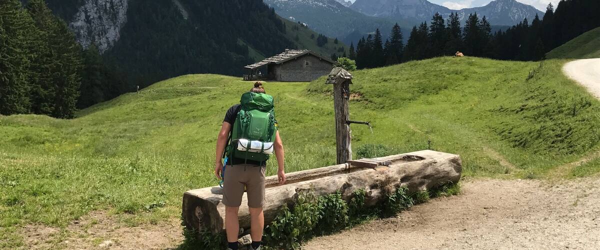 A hiker with a green backpack standing on a dirt path in a mountainous area with a wooden structure and a clear sky.