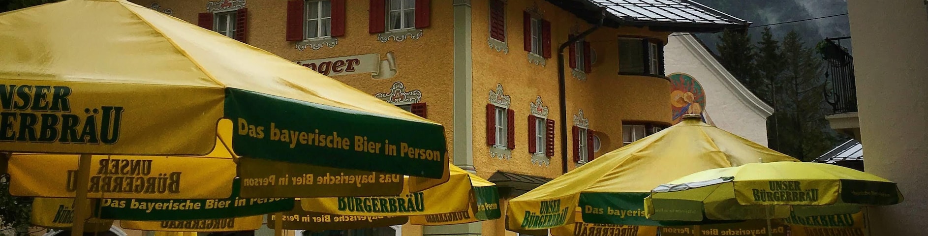 Un restaurant avec une terrasse et des parasols, une église dotée d’un clocher et une montagne en arrière-plan.
