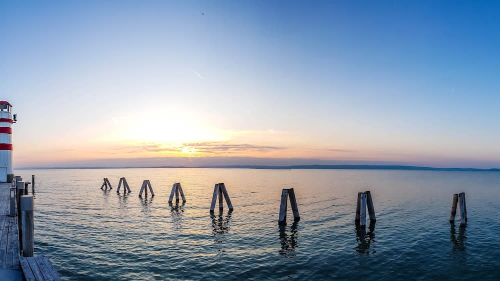 White and red striped lighthouse in Neusiedlersee, on Austrian-Hungarian border. Sun sets over the horizon in the clouds. Sky is orange, lake surface is soft and tender. Few clouds on the sky.