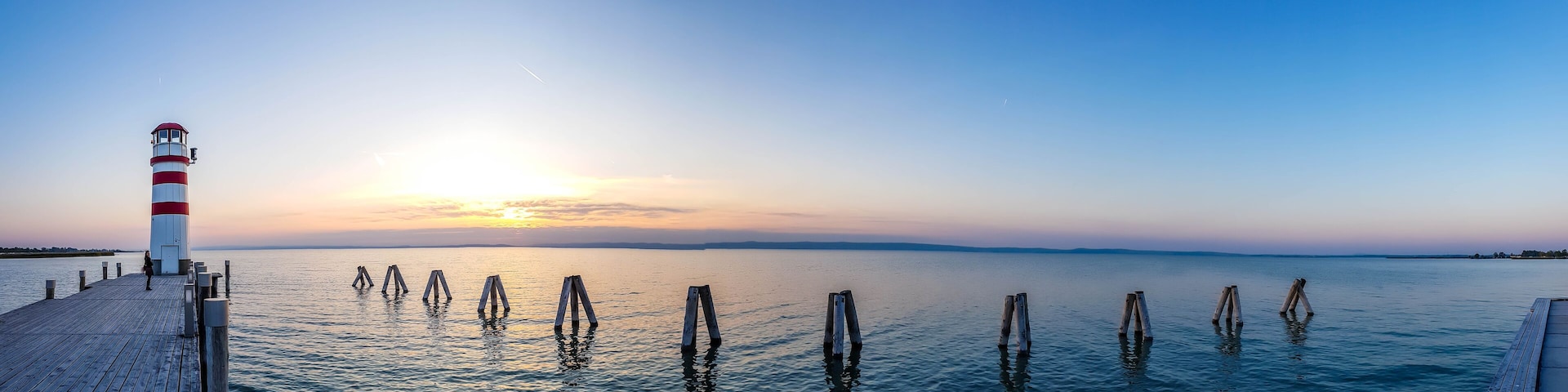 White and red striped lighthouse in Neusiedlersee, on Austrian-Hungarian border. Sun sets over the horizon in the clouds. Sky is orange, lake surface is soft and tender. Few clouds on the sky.