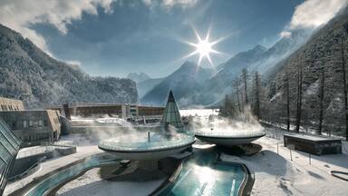 Un complejo turístico en la montaña cubierto de nieve, con un spa de aguas termales, un edificio con techo de vidrio y un cielo azul despejado.