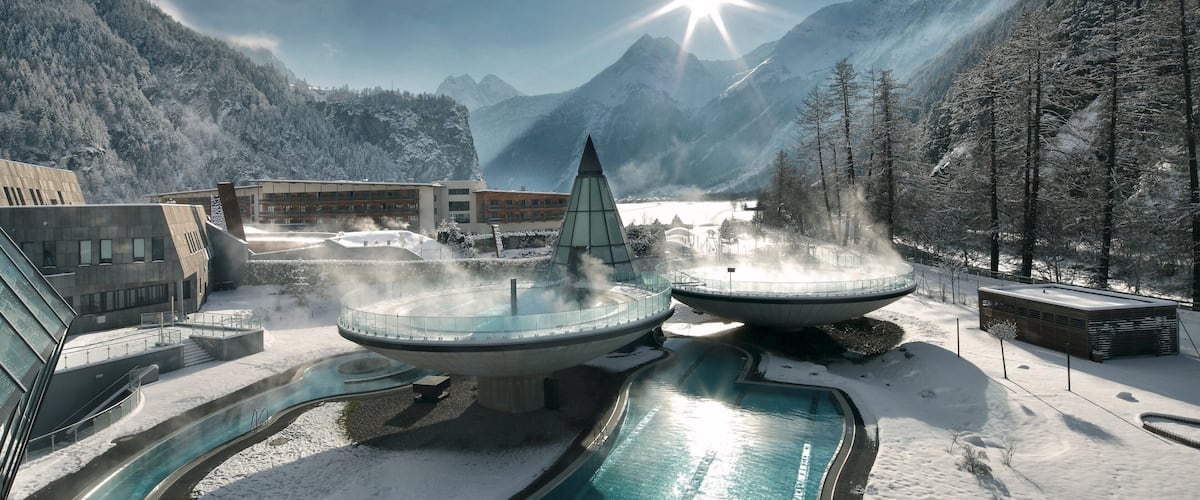 A snow-covered mountain resort with a hot spring spa, a glass-roofed building, and a clear blue sky.