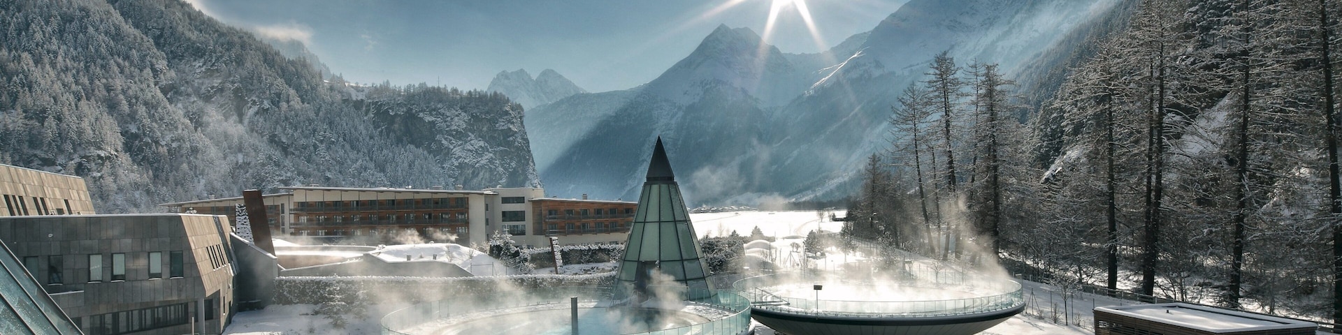 A snow-covered mountain resort with a hot spring spa, a glass-roofed building, and a clear blue sky.