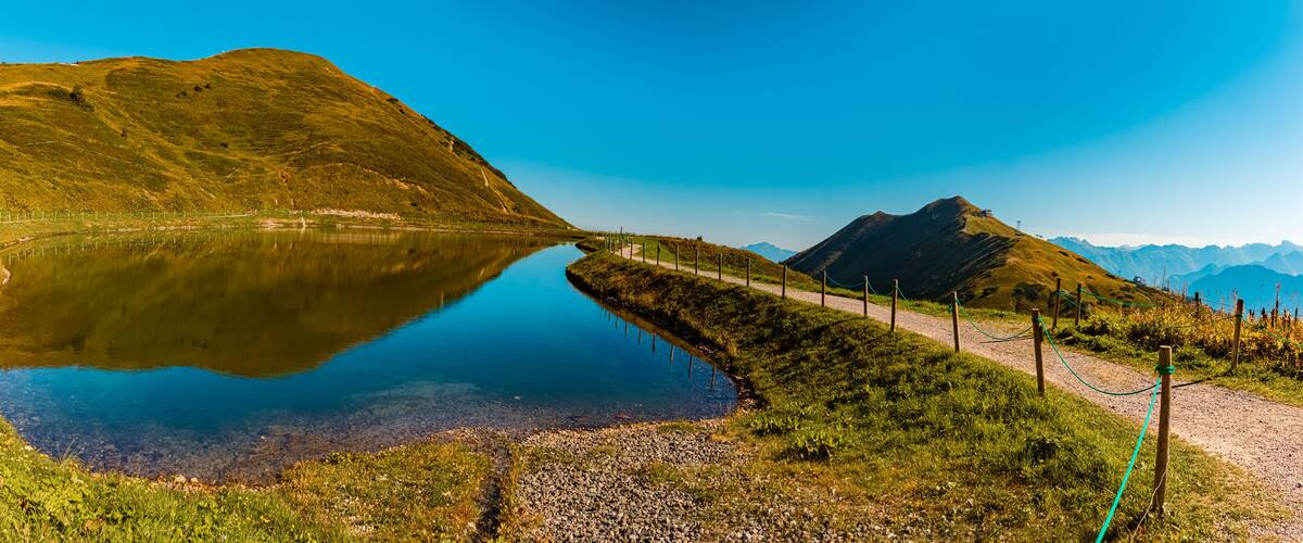 A mountain lake with a clear reflection, surrounded by grassy hills and a dirt path.