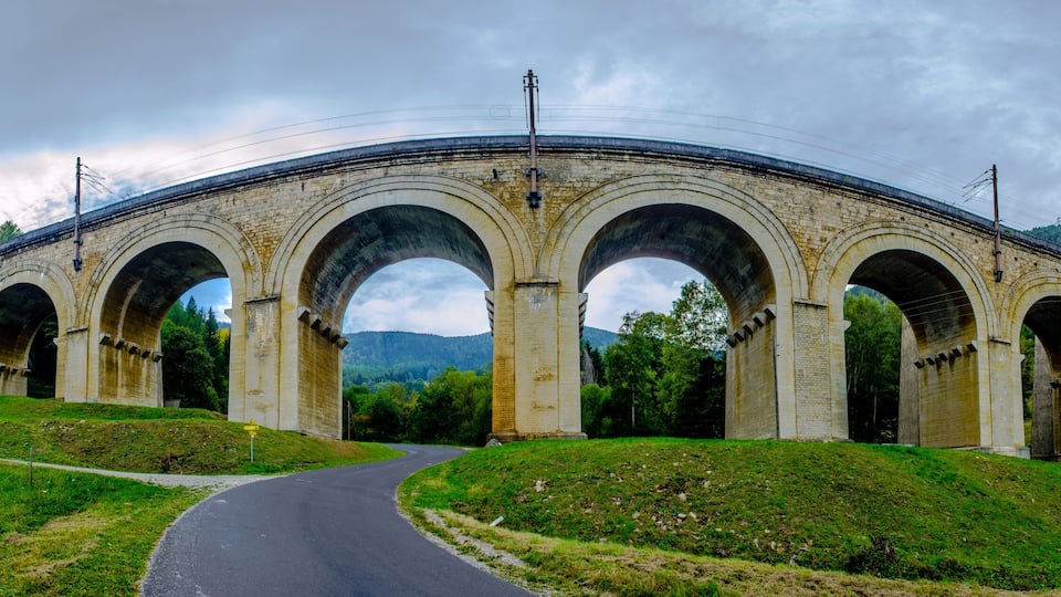 A stone viaduct with multiple arches over a road, surrounded by green hills and trees.