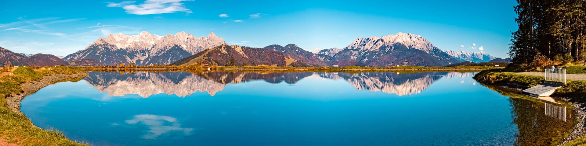 A mountain lake with clear reflections of the peaks, surrounded by greenery and a paved path.
