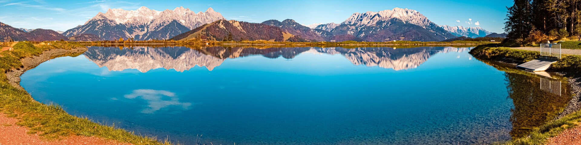 Un lago de montaña con reflejos nítidos de las cumbres, rodeado de vegetación y un camino pavimentado.