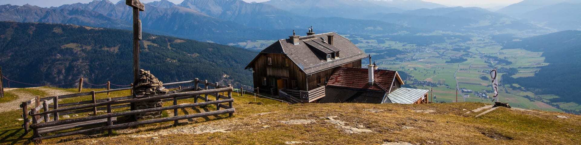A wooden cabin with a stone cross and a wooden fence on a mountain ridge.
