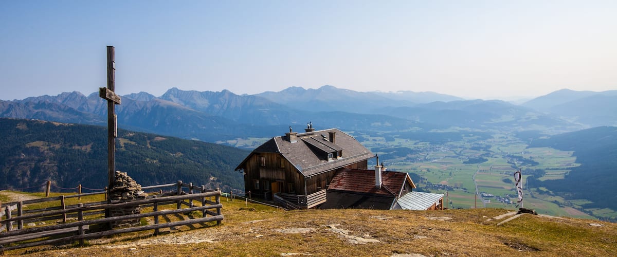 A wooden cabin with a stone cross and a wooden fence on a mountain ridge.