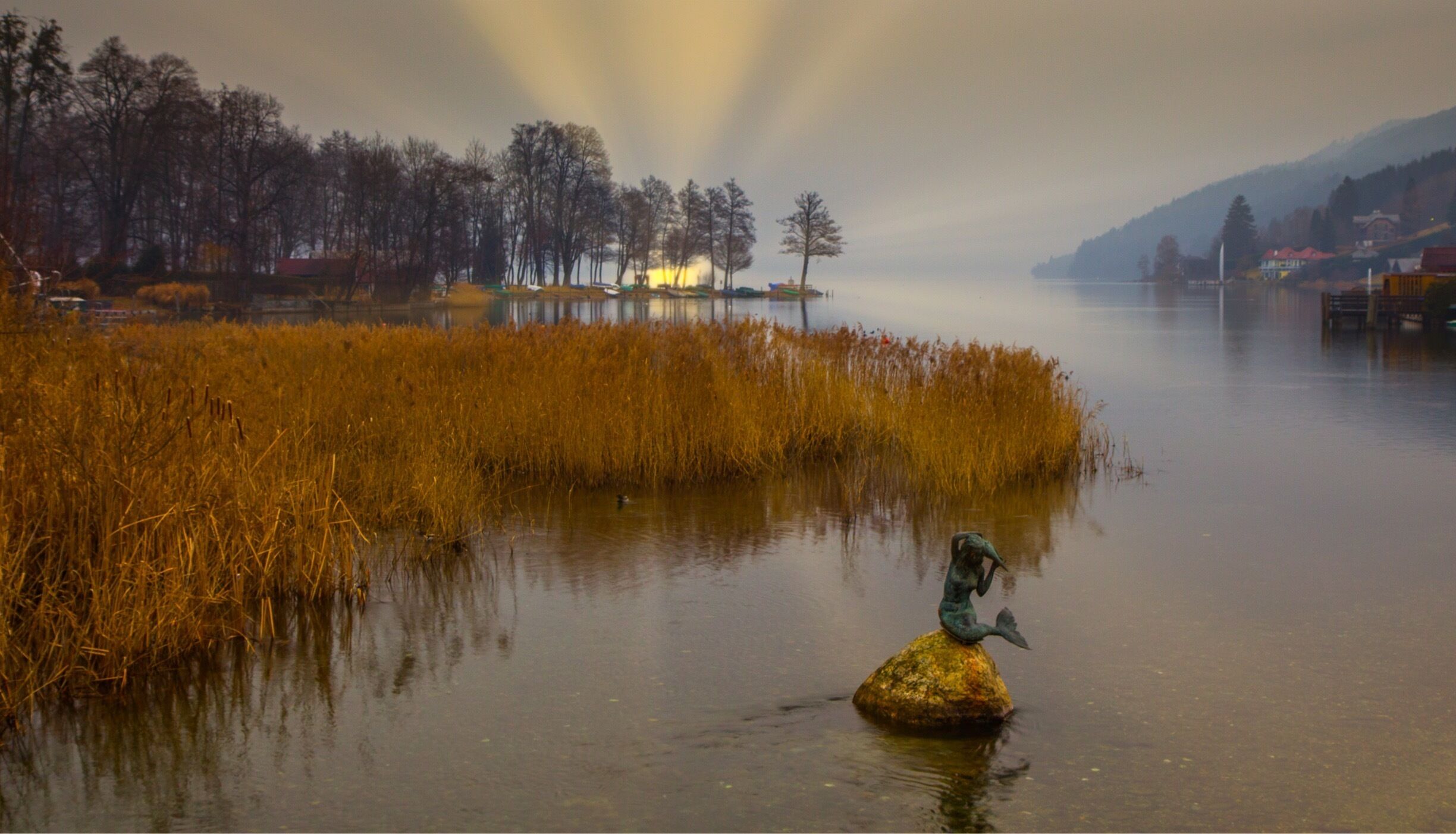 A serene lake with golden reeds, a statue of a mermaid on a rock, and a misty mountain backdrop.