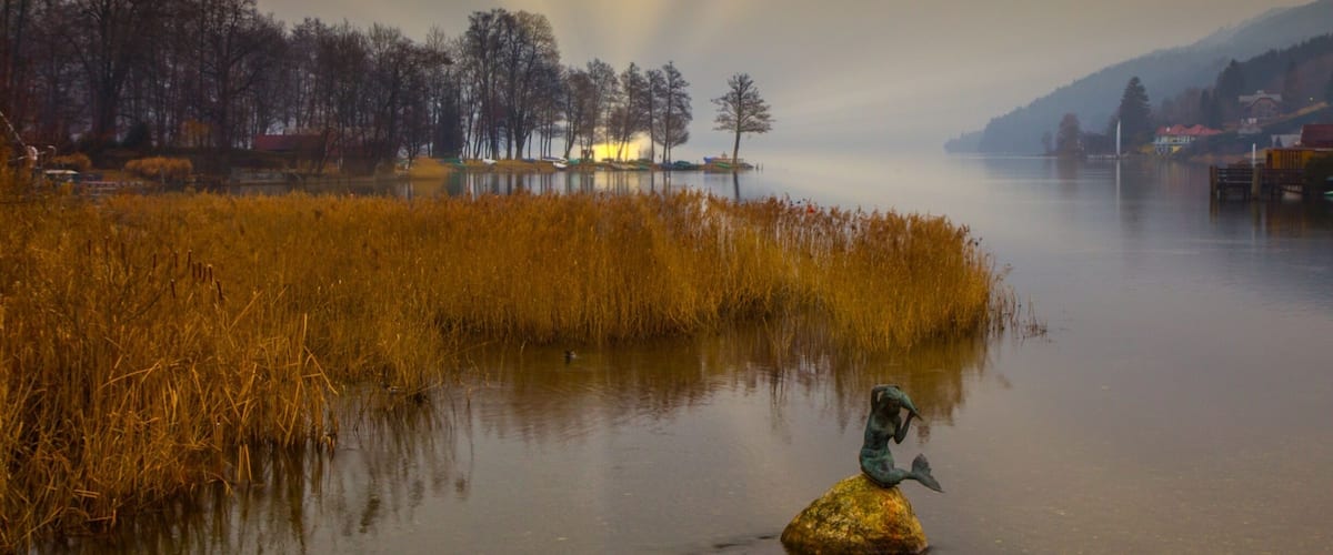 A serene lake with golden reeds, a statue of a mermaid on a rock, and a misty mountain backdrop.
