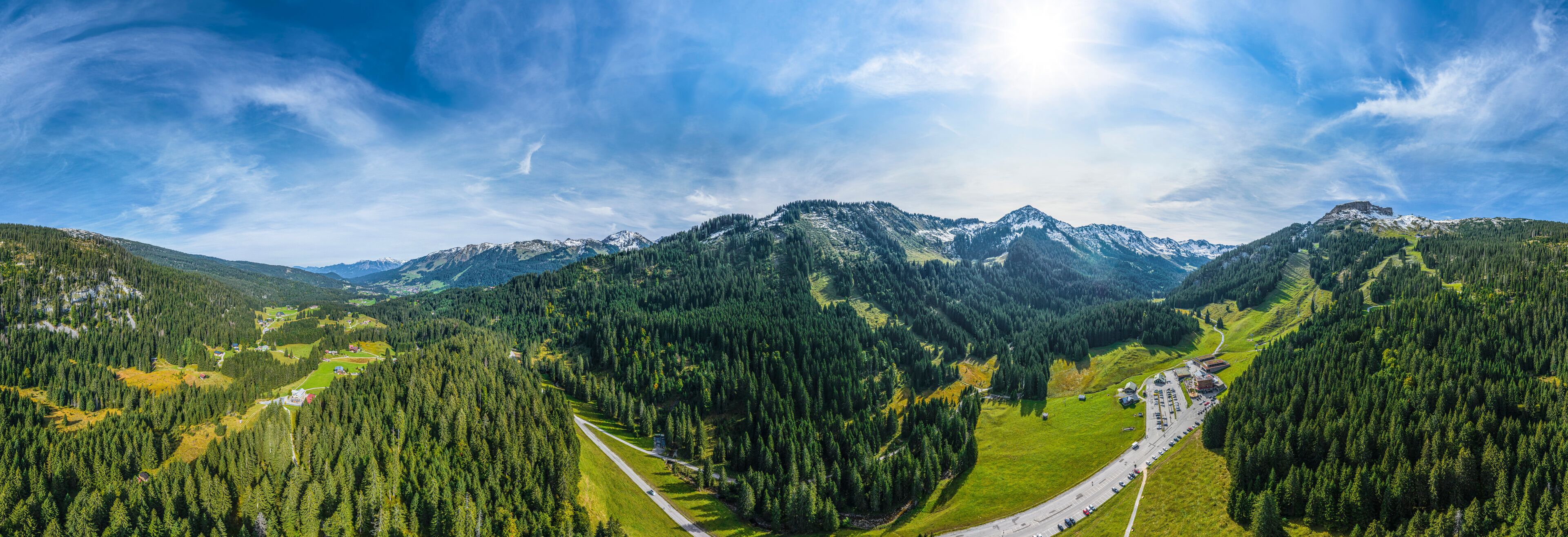 A mountainous landscape with dense forests, a winding road, and snow-capped peaks under a clear sky.