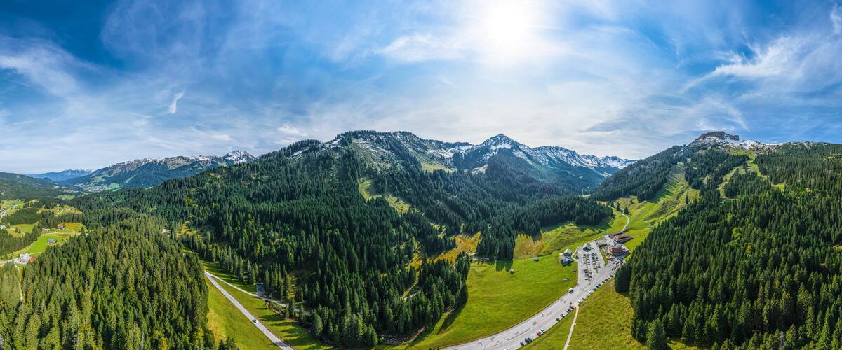 A mountainous landscape with dense forests, a winding road, and snow-capped peaks under a clear sky.