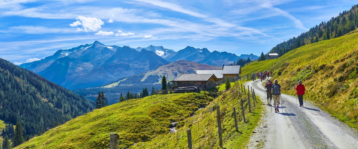 A mountain road with hikers, a wooden cabin, and snow-capped peaks.