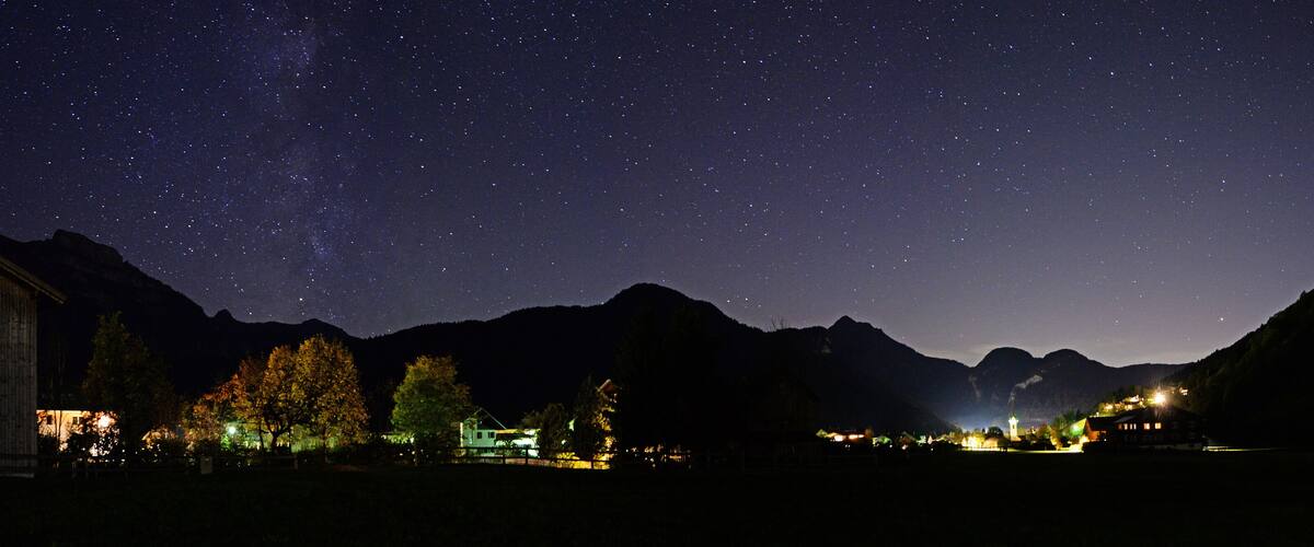 A starry night sky over a mountain town with illuminated buildings and trees.