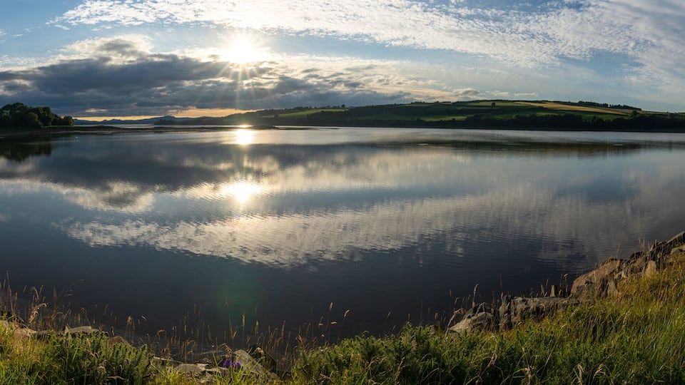 A calm lake with a path along its edge, surrounded by greenery and hills in the background.