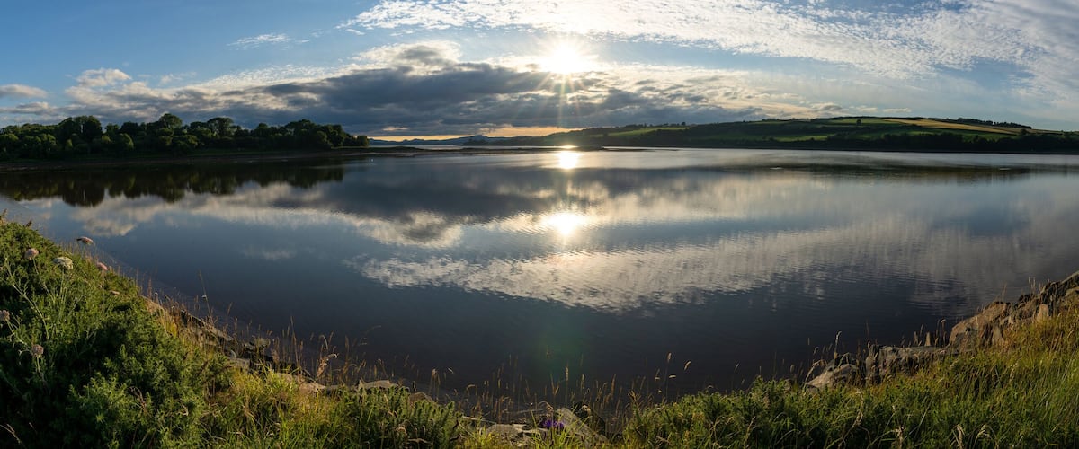 A calm lake with a path along its edge, surrounded by greenery and hills in the background.