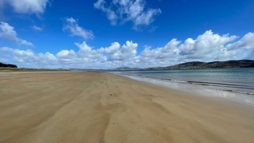 A wide sandy beach with gentle waves, a clear blue sky, and distant hills.