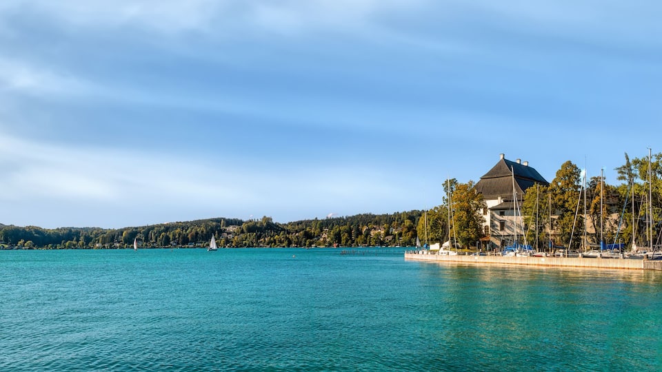 A calm lake with sailboats, a dock with boats, and a building with a green roof.