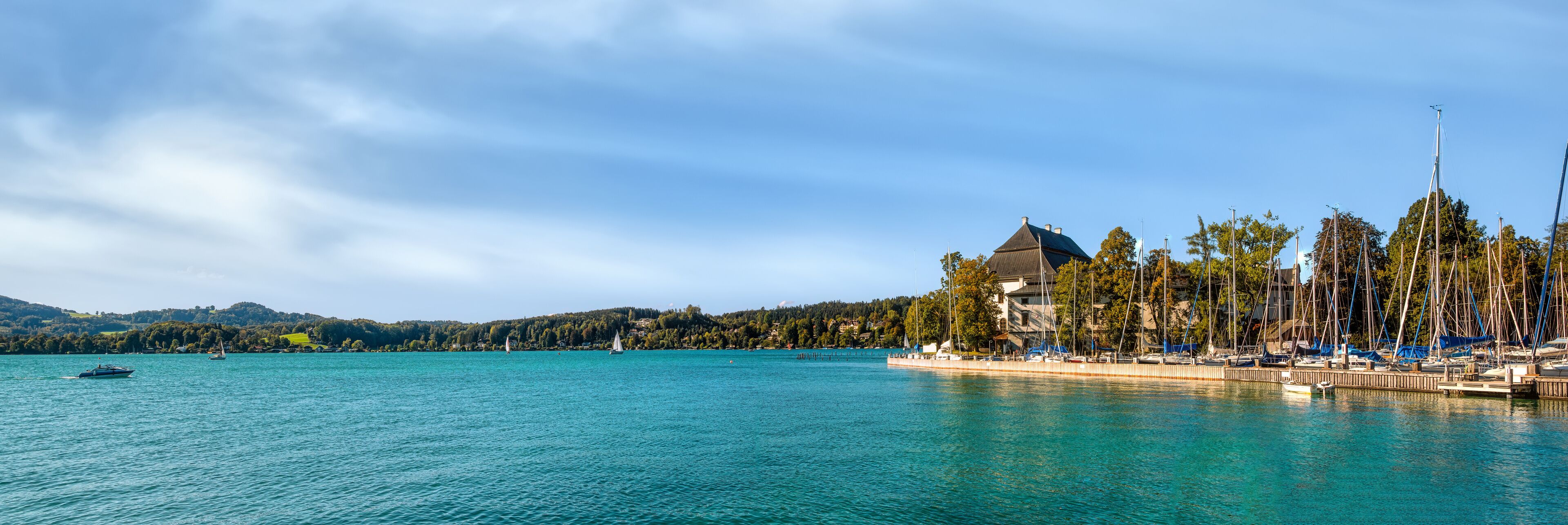 A calm lake with sailboats, a dock with boats, and a building with a green roof.