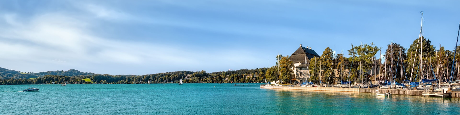 A calm lake with sailboats, a dock with boats, and a building with a green roof.