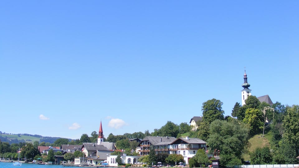 A lakeside town with a prominent church steeple, residential buildings, and lush greenery.