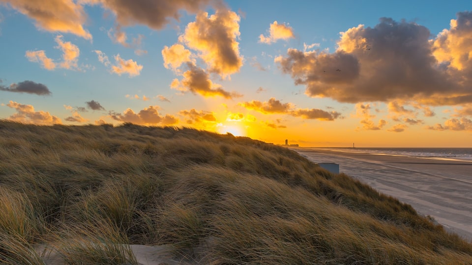 A sunset over a beach with dunes and a distant lighthouse.