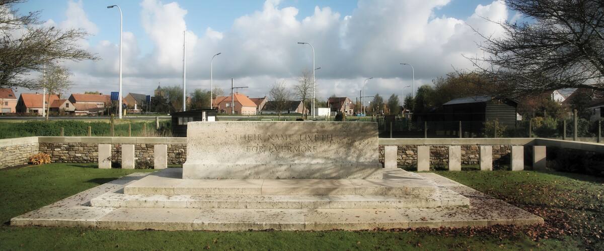 A memorial with inscriptions, surrounded by a grassy area, trees, and houses in the background.