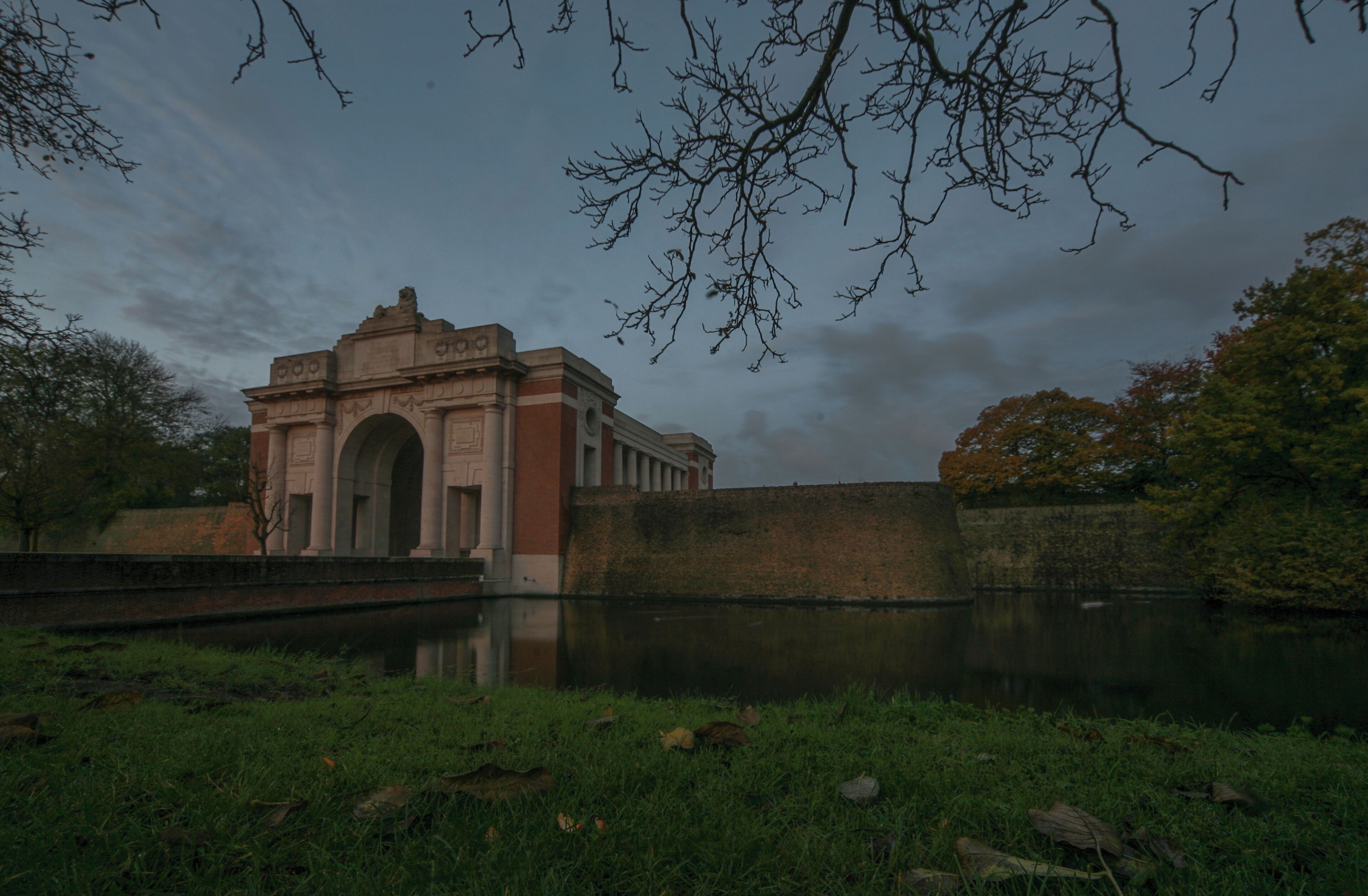 A historic arch bridge over a calm river, surrounded by greenery and a cloudy sky.