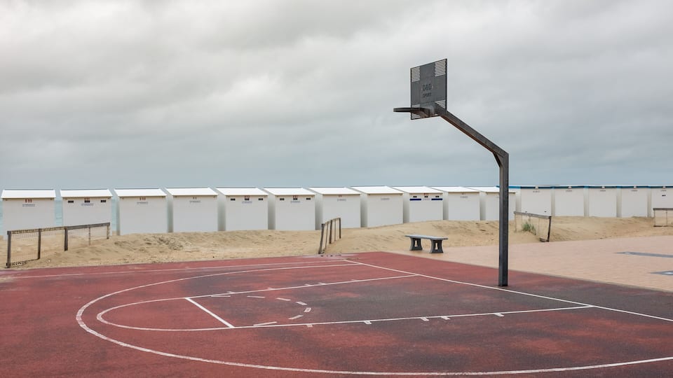 An outdoor basketball court with a red surface, a bench, and a wall of numbered lockers.