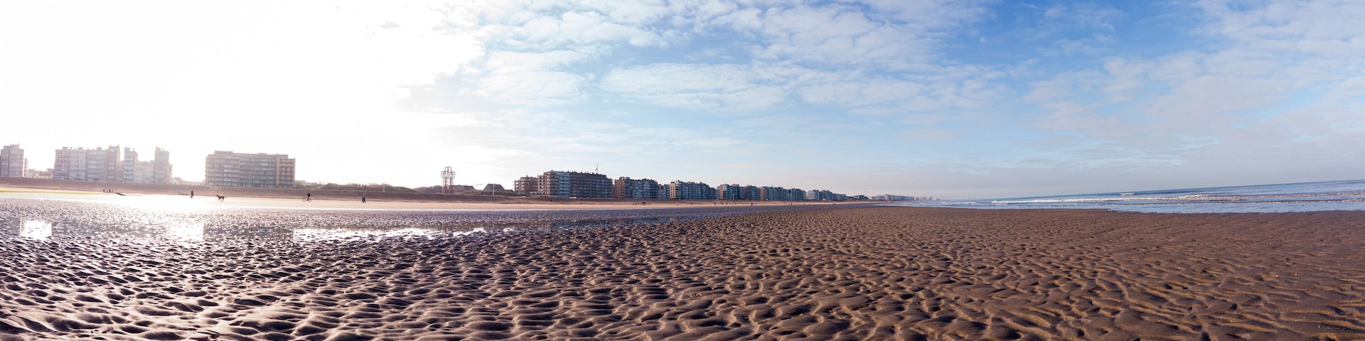 A panoramic view of a vast sandy beach with a city skyline under a bright blue sky. The tranquil scene evokes peacefulness and escape, perfect for a background or travel concept.