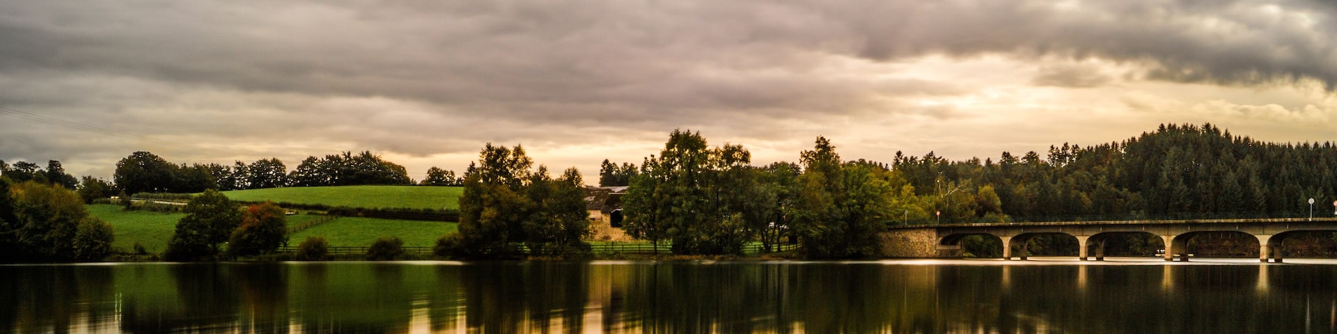 A calm lake with a stone bridge spanning across it, surrounded by trees and grassy fields under a cloudy sky.