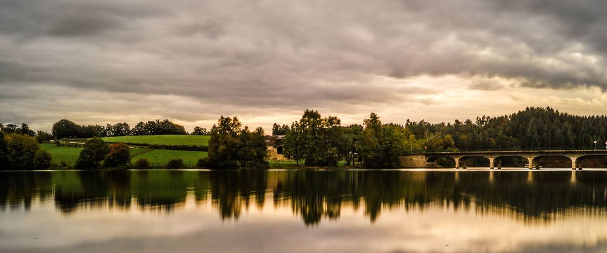 A calm lake with a stone bridge spanning across it, surrounded by trees and grassy fields under a cloudy sky.
