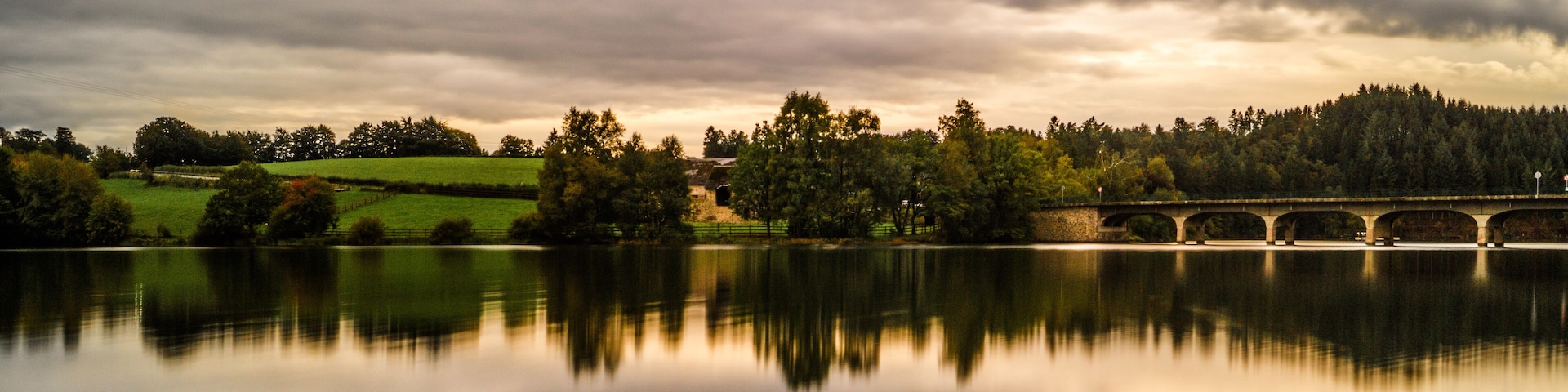 A calm lake with a stone bridge spanning across it, surrounded by trees and grassy fields under a cloudy sky.