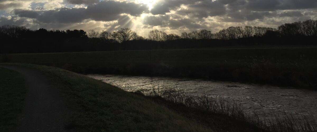 A river with a path alongside it, under a cloudy sky with sunlight breaking through.